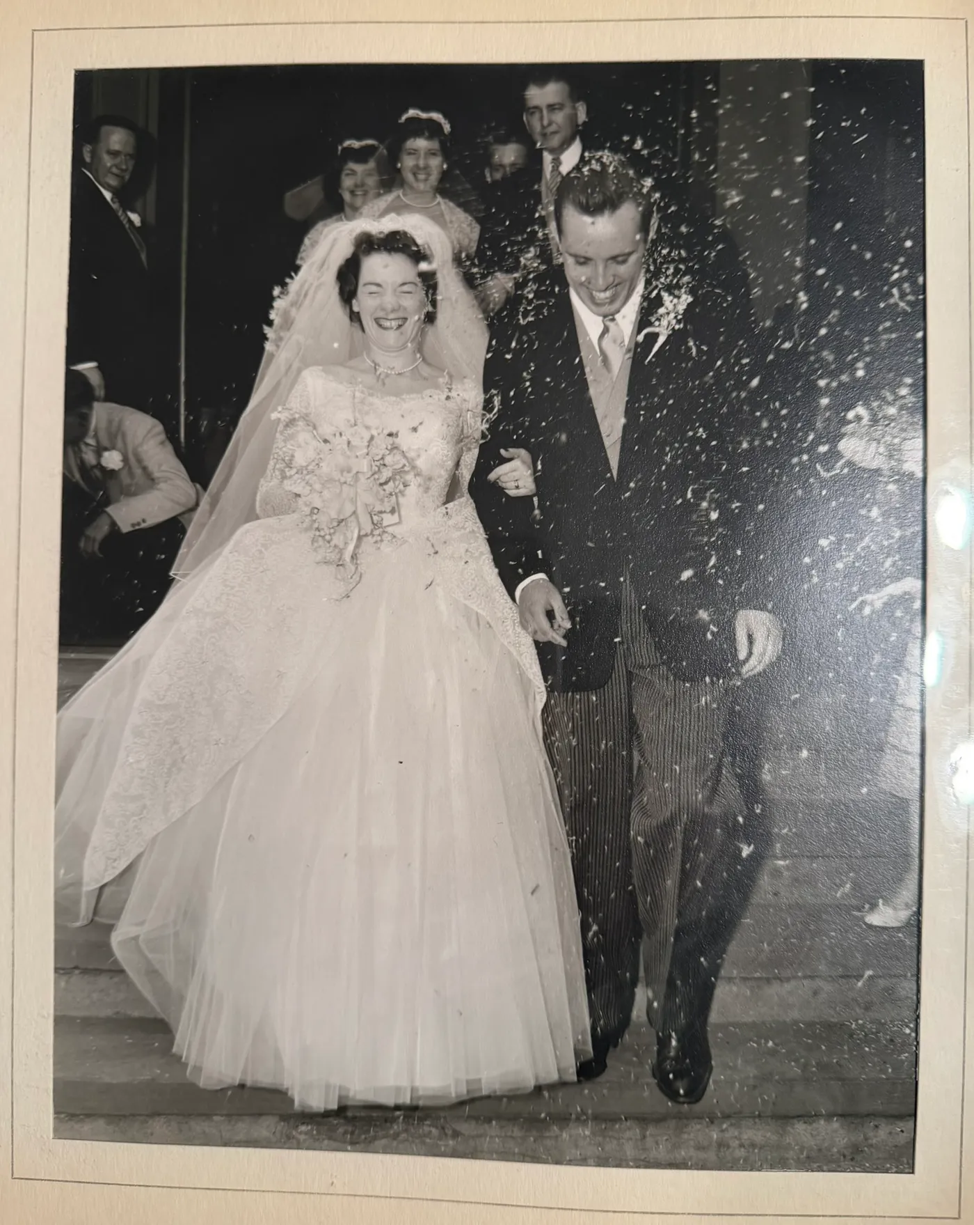 A black-and-white wedding photograph of a smiling bride and groom walking through a shower of rice while guests look on
